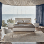 DOLLY bed designed by DANIELE  LO SCALZO MOSCHERI. DOLLY puts an original slant on the traditional headboard which envelops the edges of the bed from the bottom, swathing it like an elegant shell and uniting the two separate cushions. Available with three versions of the feet in wood and one in metal with the option of choosing the look and height from the ground that suits the customer best. The bed is designed for a fixed bed base or container unit. Upholstered in fabric or leather with the option of choosing from a variety of materials and finishes for the cushions and external shell of the headboard. DOLLY  fuses contemporary cool and an artisanal aesthetic which brings its sartorial stylishness to the fore.