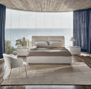 DOLLY bed designed by DANIELE  LO SCALZO MOSCHERI. DOLLY puts an original slant on the traditional headboard which envelops the edges of the bed from the bottom, swathing it like an elegant shell and uniting the two separate cushions. Available with three versions of the feet in wood and one in metal with the option of choosing the look and height from the ground that suits the customer best. The bed is designed for a fixed bed base or container unit. Upholstered in fabric or leather with the option of choosing from a variety of materials and finishes for the cushions and external shell of the headboard. DOLLY  fuses contemporary cool and an artisanal aesthetic which brings its sartorial stylishness to the fore.