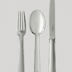 NORMANDIE cutlery collection. Linking Le Havre to New York, the famous ocean liner Normandie embodied Art Deco and its French artists and was honored at the 1925 Exposition Universelle. This timeless cutlery, designed by JEAN PUIFORCAT in 1934, graced the tables of the dining rooms in first-class suites. Now available in silver-plated metal, the elegant silhouette, with its smooth surfaces and delicate curves accentuated by an ornamental disk, is also offered in a line of table accessories.