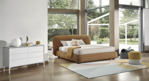 NOA bed designed by DANIELE  LO SCALZO MOSCHERI. NOA is a sculptured bed with a harmoniously rational structure drawing inspiration from rocks sculpted by the wind. Soft and voluminous, the simple-looking headboard has a three-dimensional beauty which is accentuated further by the topstitching on the curved surfaces. The item is available with three versions of the feet in wood or one version of the feet in metal with the option of choosing a look and height from the ground that suits the customer best. NOA is available with a fixed bed base or container unit. Can be upholstered in fabric or leather.