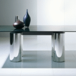 Table SERENISSIMO by LELLA  and MASSIMO VIGNELLI. The precisely delineated SERENISSIMO renders a table as a graphic composition, with four columns and a linear steel girder to float the table top above. In the key of LELLA and MASSIMO VIGNELLI‚ÄôS original design, the creation speaks through its geometry and materials. With its elemental shapes defined by refined metallic finishes or soft tones of venetian stucco, and by a vast glass top ‚Äî up to three meters long ‚Äî extended atop the altar-like base, for a harmony composed of monumental proportions.