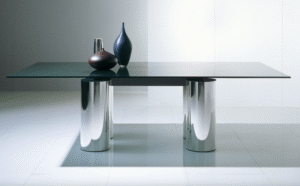 Table SERENISSIMO by LELLA  and MASSIMO VIGNELLI. The precisely delineated SERENISSIMO renders a table as a graphic composition, with four columns and a linear steel girder to float the table top above. In the key of LELLA and MASSIMO VIGNELLI‚ÄôS original design, the creation speaks through its geometry and materials. With its elemental shapes defined by refined metallic finishes or soft tones of venetian stucco, and by a vast glass top ‚Äî up to three meters long ‚Äî extended atop the altar-like base, for a harmony composed of monumental proportions.