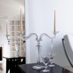 MILLE-NUITS candelabra. The bevel-cut arms of the candelabra, amidst which stand two decorative crystal swirls, glitter luminously. Each candelabra arm is adorned with dangling arrowhead-shaped crystals that catch the light in a scintillating manner. Candles nestle within crystal candleholders shaped like blooming daffodils. The MILLE NUITS candelabra is a sumptuous item, made in two-, three-, or five-light versions; also available in gold. Hurricane shades, available separately, emphasize every candle flicker and intensify the luminosity. Designed for Baccarat by MATHIAS, MILLE NUITS is a reference to the 1001 Tales of The Arabian Nights. The MILLE NUITS collection also encompasses chandeliers, stemware, and home d√©cor.